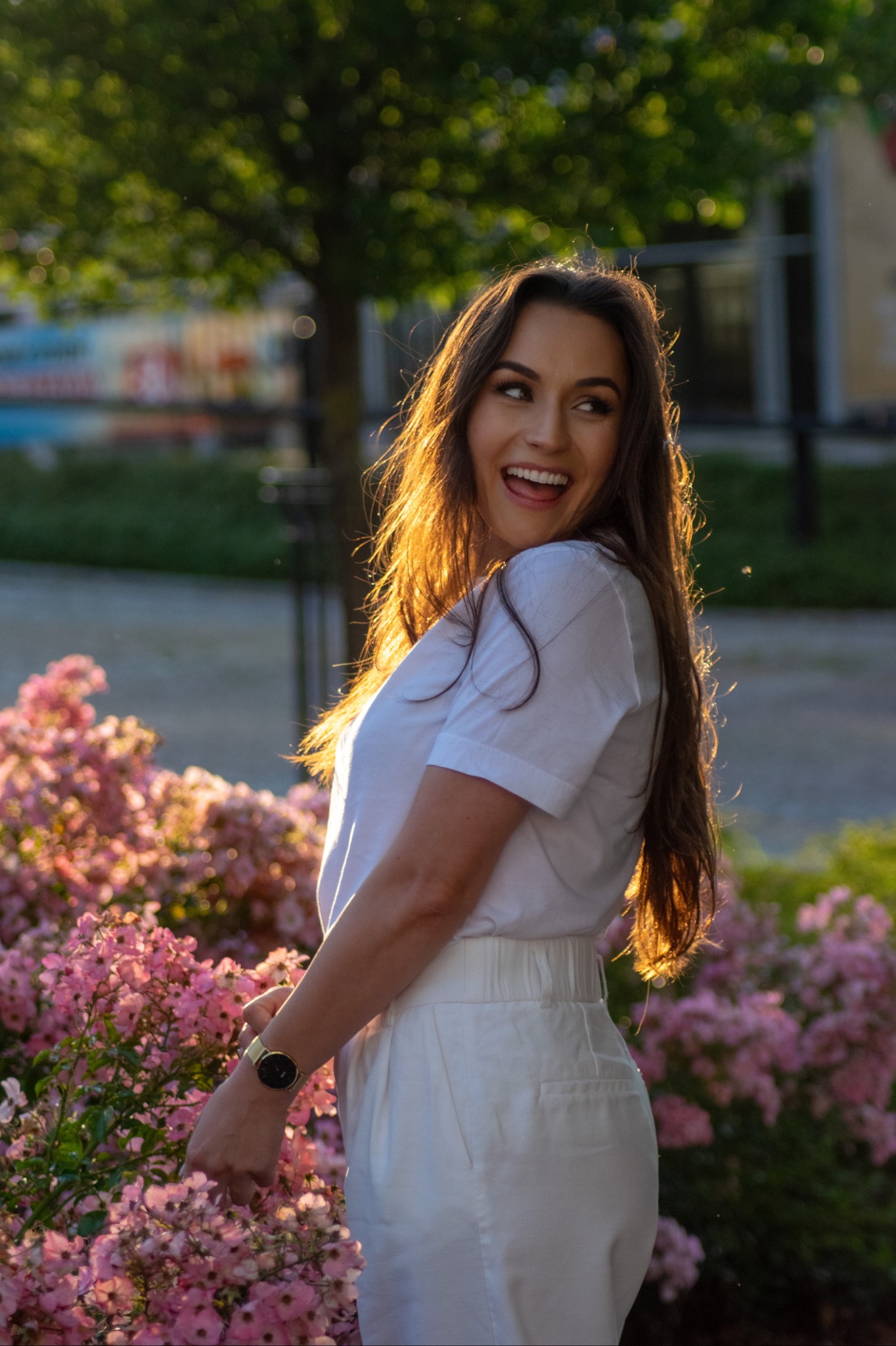 Uśmiechnięta brunetka w białym stroju, z zegarkiem na ręce, pozuje przy kwitnącym krzewie róż w promieniach zachodzącego słońca, odwrócona w bok.