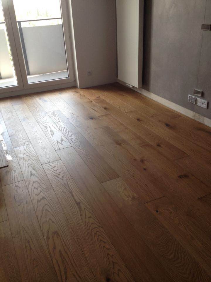 Ułożona, lakierowana podłoga z desek w nowym mieszkaniu, widoczne gniazdka elektryczne przy ścianie i okno balkonowe.