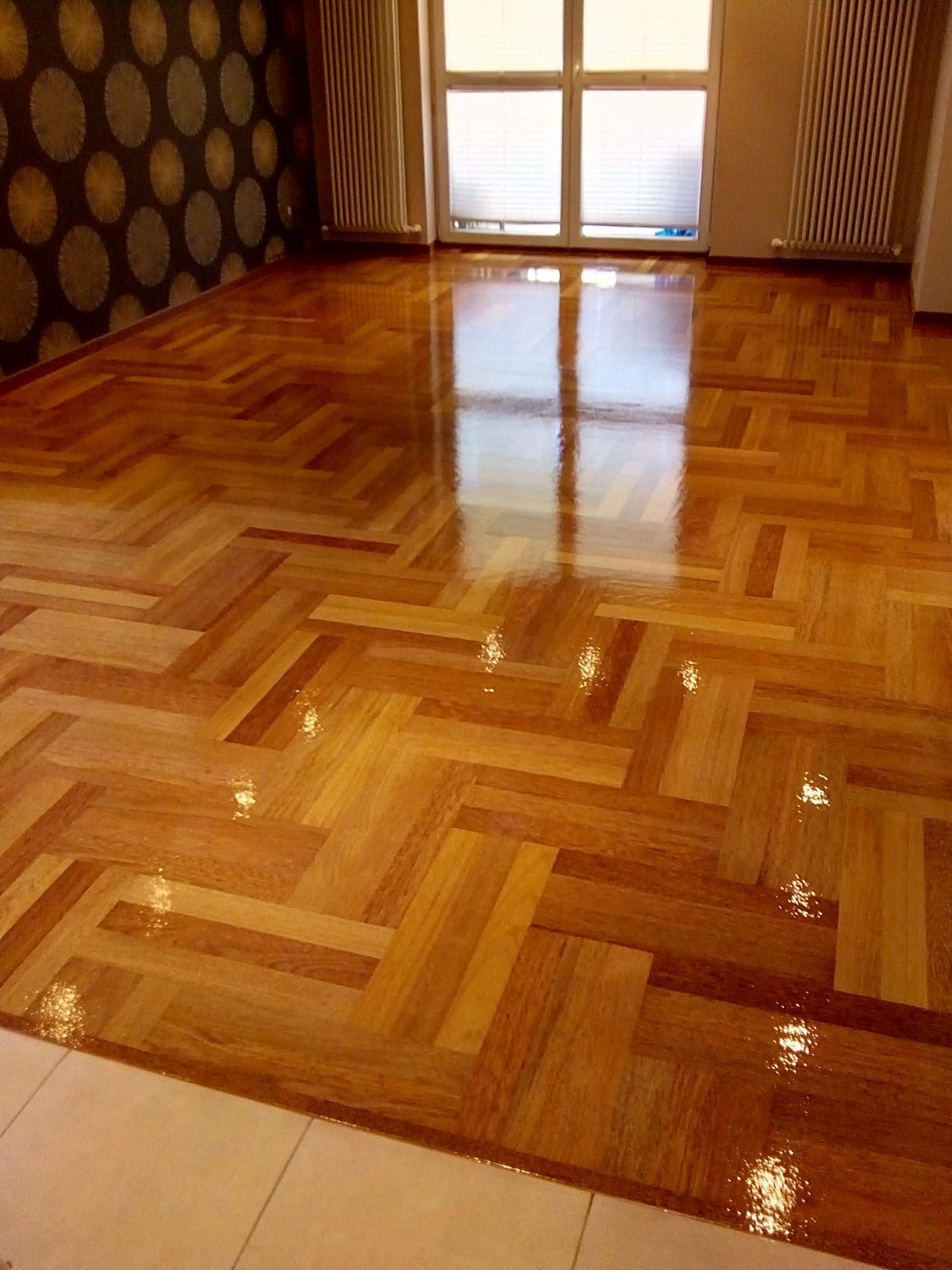 Błyszczący parkiet ułożony we wzór jodełki, odbijający światło z okna, na tle ściany z wzorzystą tapetą i grzejnikami.