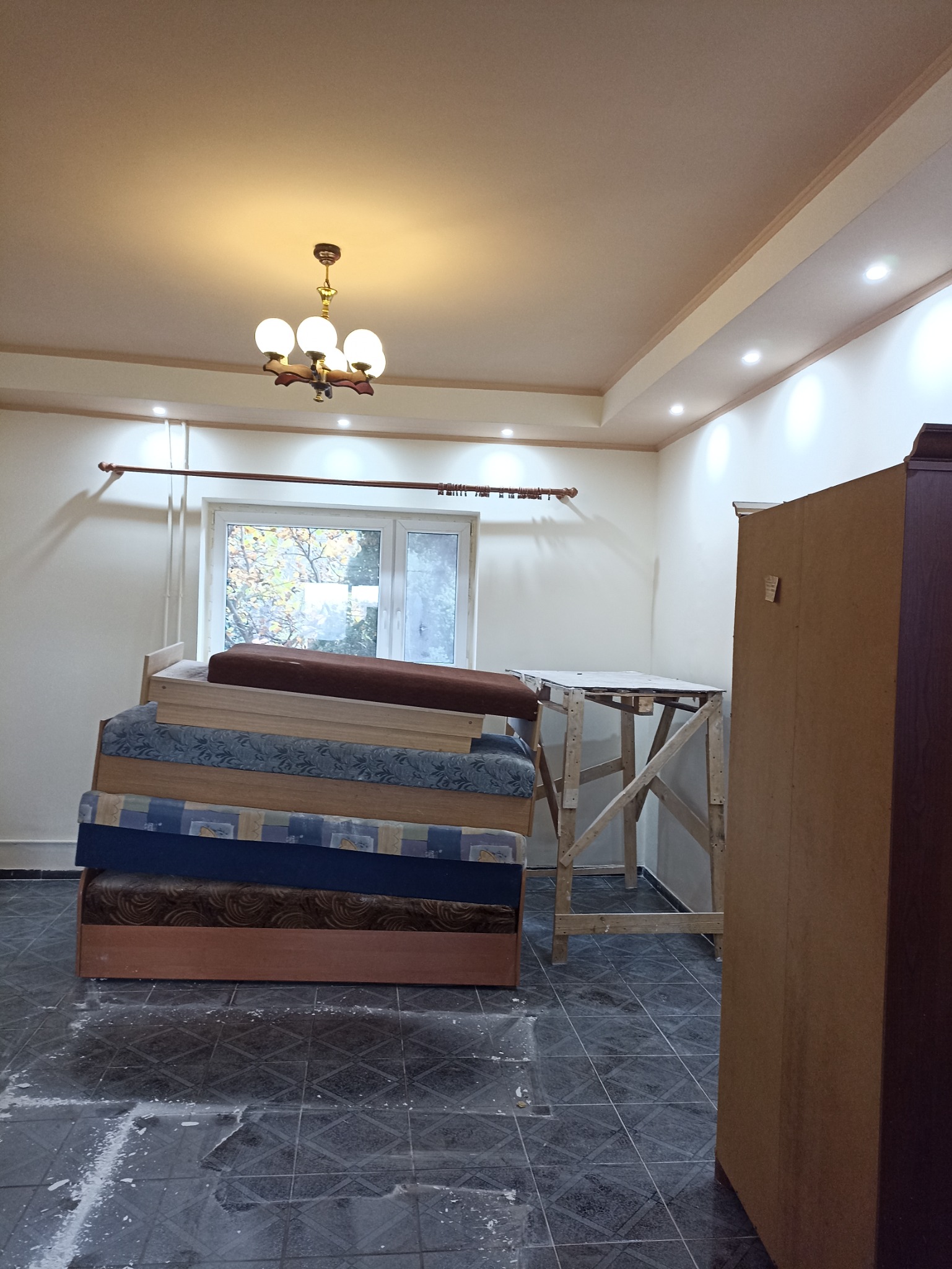 Pokój w trakcie remontu: widoczny stos materacy, drewniany stół roboczy, ślady gipsu na podłodze z płytek, sufit z oświetleniem halogenowym i lampą wiszącą.