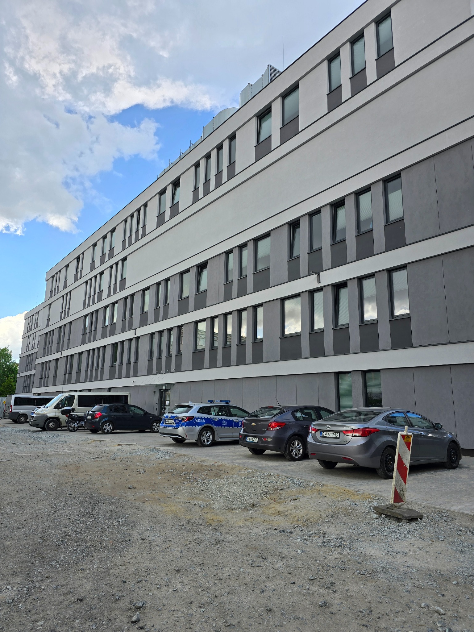 Nowoczesny budynek Komisariatu Policji we Wrocławiu z szarą elewacją i rzędami okien, zaparkowane samochody służbowe na żwirowym parkingu.