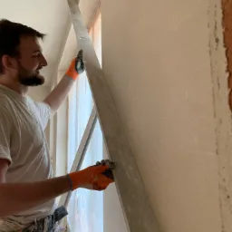 Pracownik w pomarańczowych rękawicach wygładza ścianę długą łatą, na ścianie widoczne ślady po nałożonym tynku.