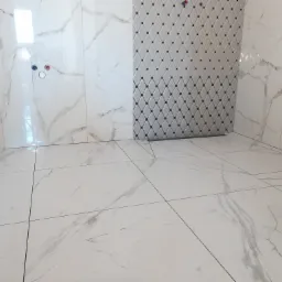 Wykończona łazienka z białymi, marmurkowymi płytkami na ścianach i podłodze, z fragmentem ściany wyłożonym płytkami w romby z czarnymi akcentami, widoczne przyłącza wodne.