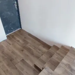 Ułożona podłoga z paneli drewnopodobnych na schodach, widoczna ciemna, brudna, pomazana drzwi wejściowe i biała ściana.