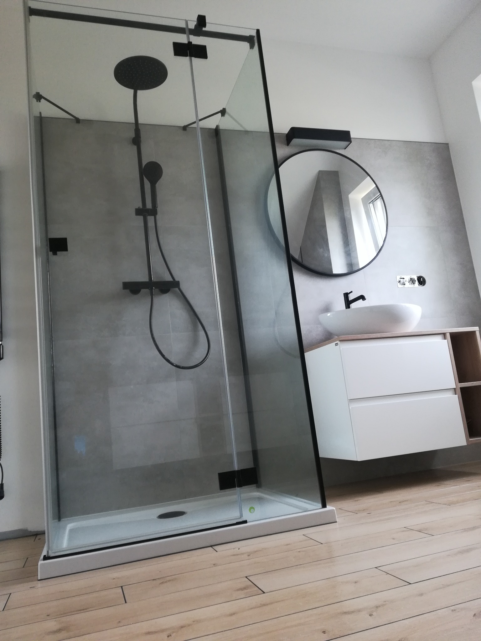Nowoczesna łazienka z kabiną prysznicową typu walk-in z czarnymi detalami, okrągłym lustrem i podwieszaną szafką z umywalką nablatową.