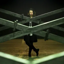 Portret kobiety w czarnym płaszczu, stojącej za metalową konstrukcją w kształcie litery X, oświetlonej jaskrawym światłem neonowym w tle.