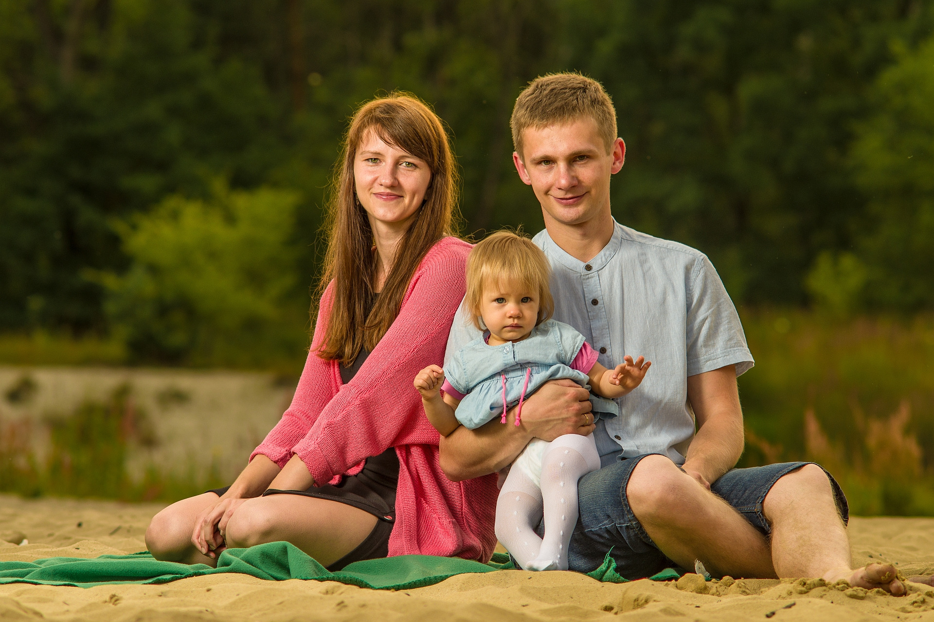 Rodzina siedząca na piasku: mama w różowym swetrze, tata w niebieskiej koszuli trzymający na kolanach córeczkę w dżinsowej sukience i białych rajstopach.