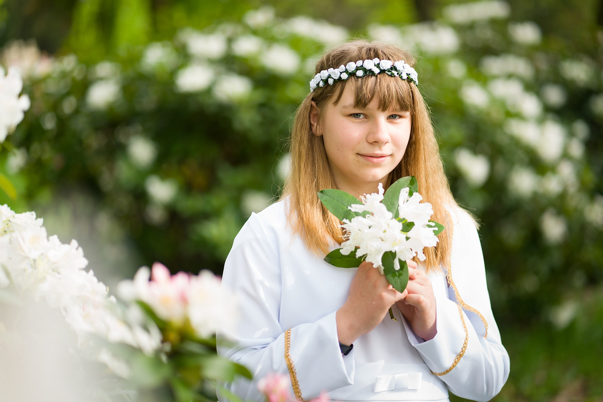 Dziewczynka w białej albie komunijnej z wiankiem z białych kwiatów na głowie, trzymająca bukiet białych kwiatów na tle rozkwitających krzewów.
