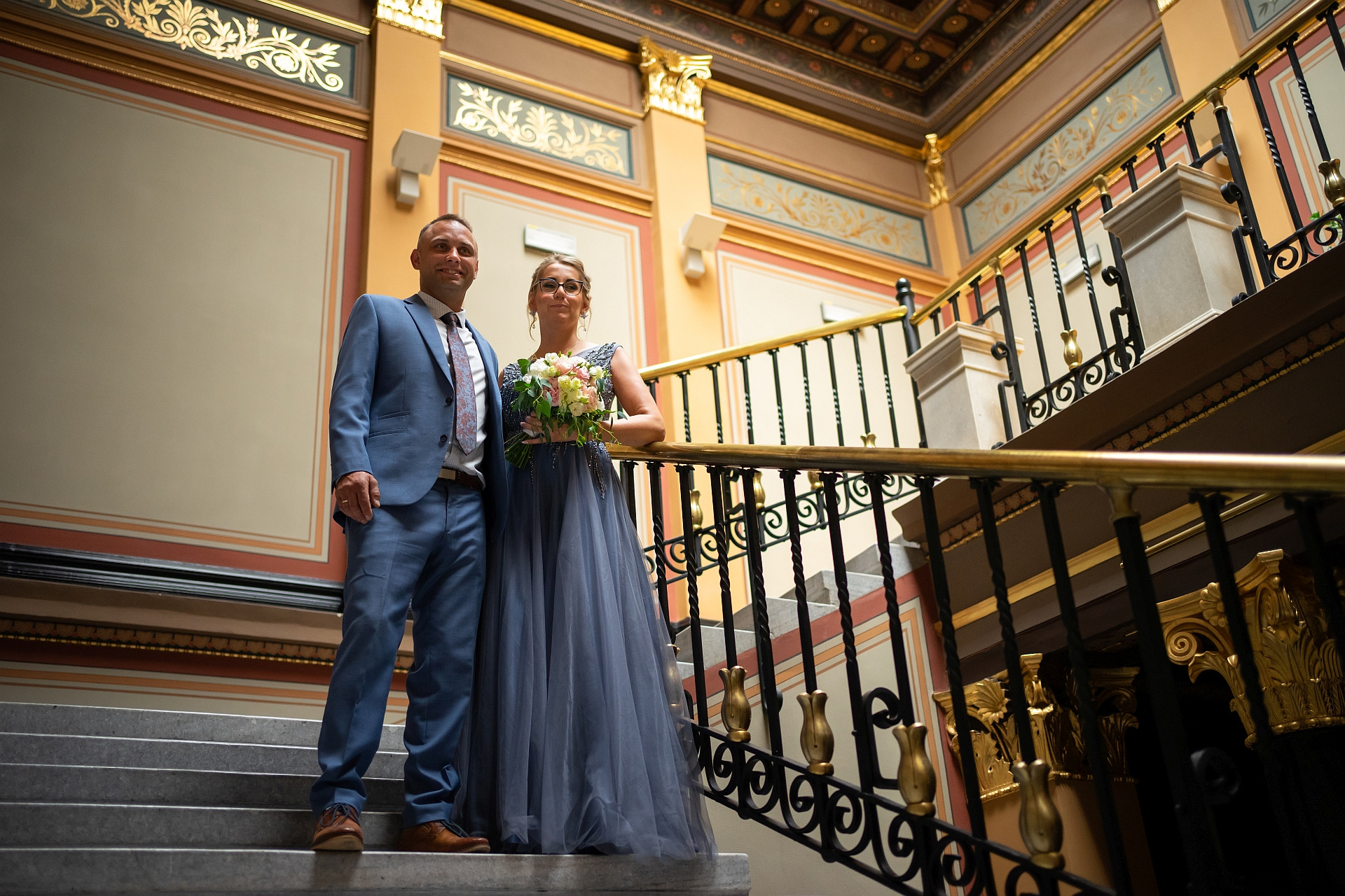 Para młoda w eleganckich strojach pozuje na schodach z ozdobnymi balustradami w Chorzowie. Panna młoda w szarej sukni trzyma bukiet kwiatów, pan młody w niebieskim garniturze.