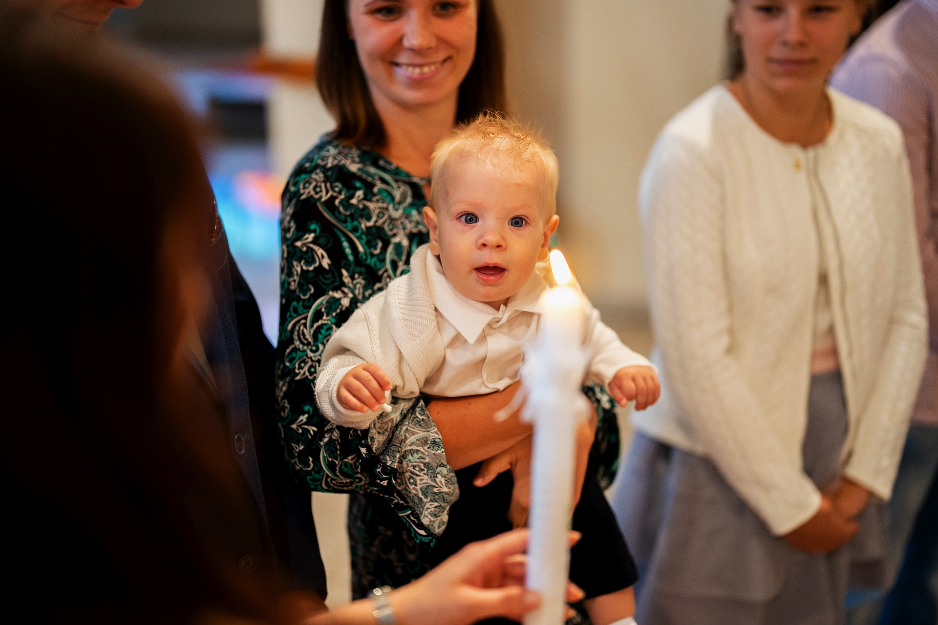 Uśmiechnięta kobieta trzyma na rękach niemowlę z rozjaśnionymi włosami w białym ubranku, w tle inne osoby i zapalona świeca.