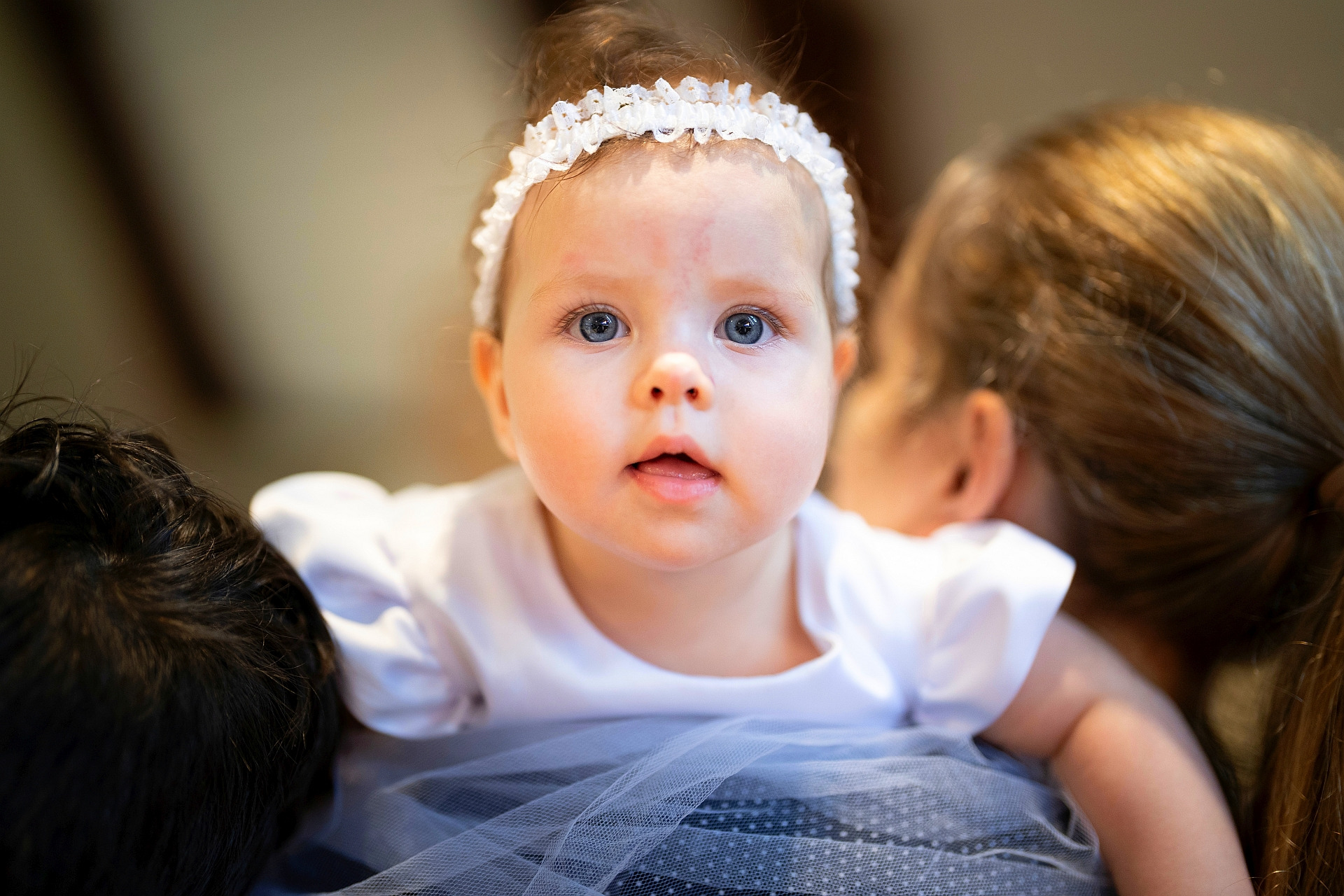 Portret niemowlęcia w białej sukience i koronkowej opasce na głowie, o dużych niebieskich oczach, opierającego się na ramieniu dorosłej osoby.