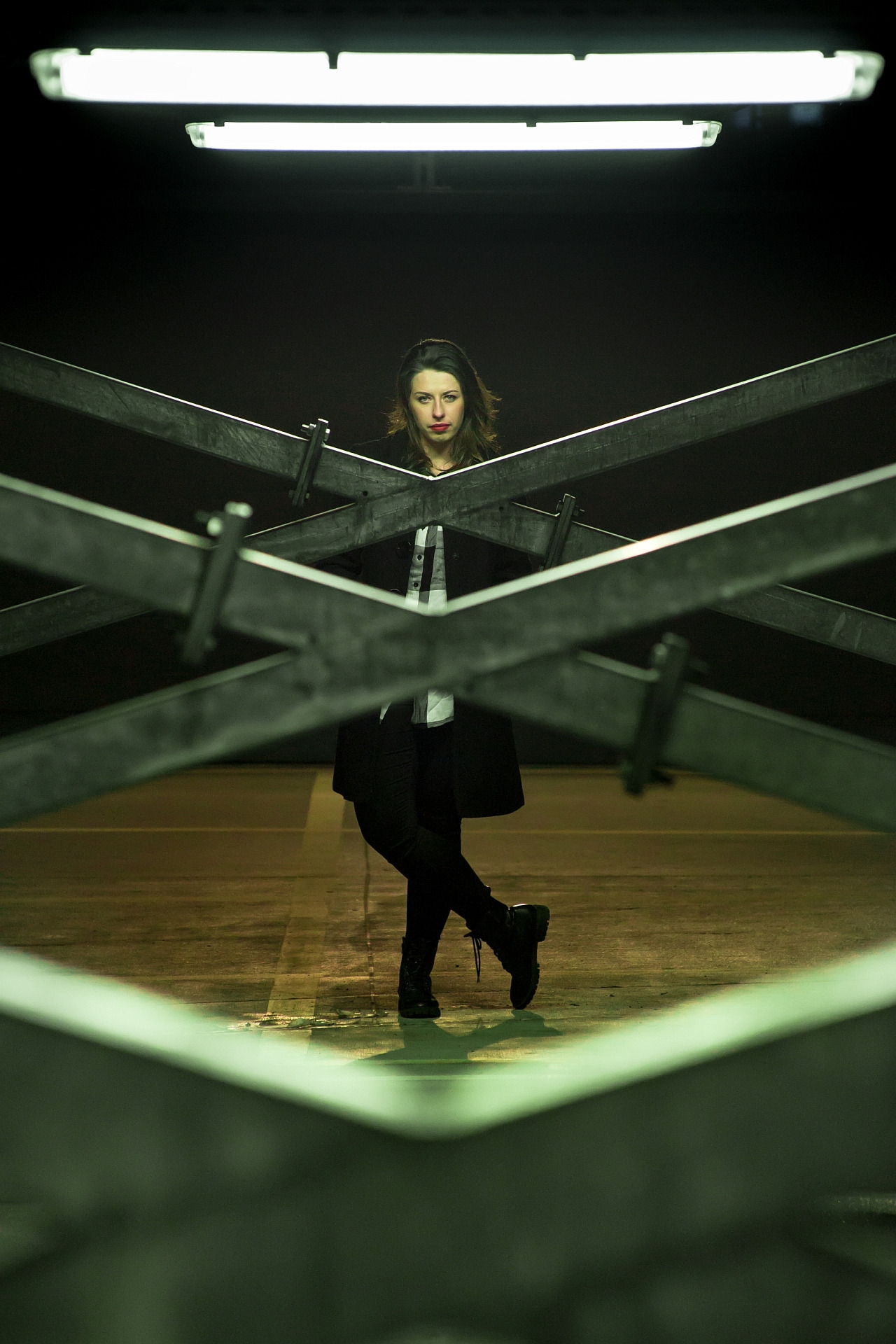 Portret kobiety w czarnym płaszczu, stojącej za metalową konstrukcją w kształcie litery X, oświetlonej jaskrawym światłem neonowym w tle.