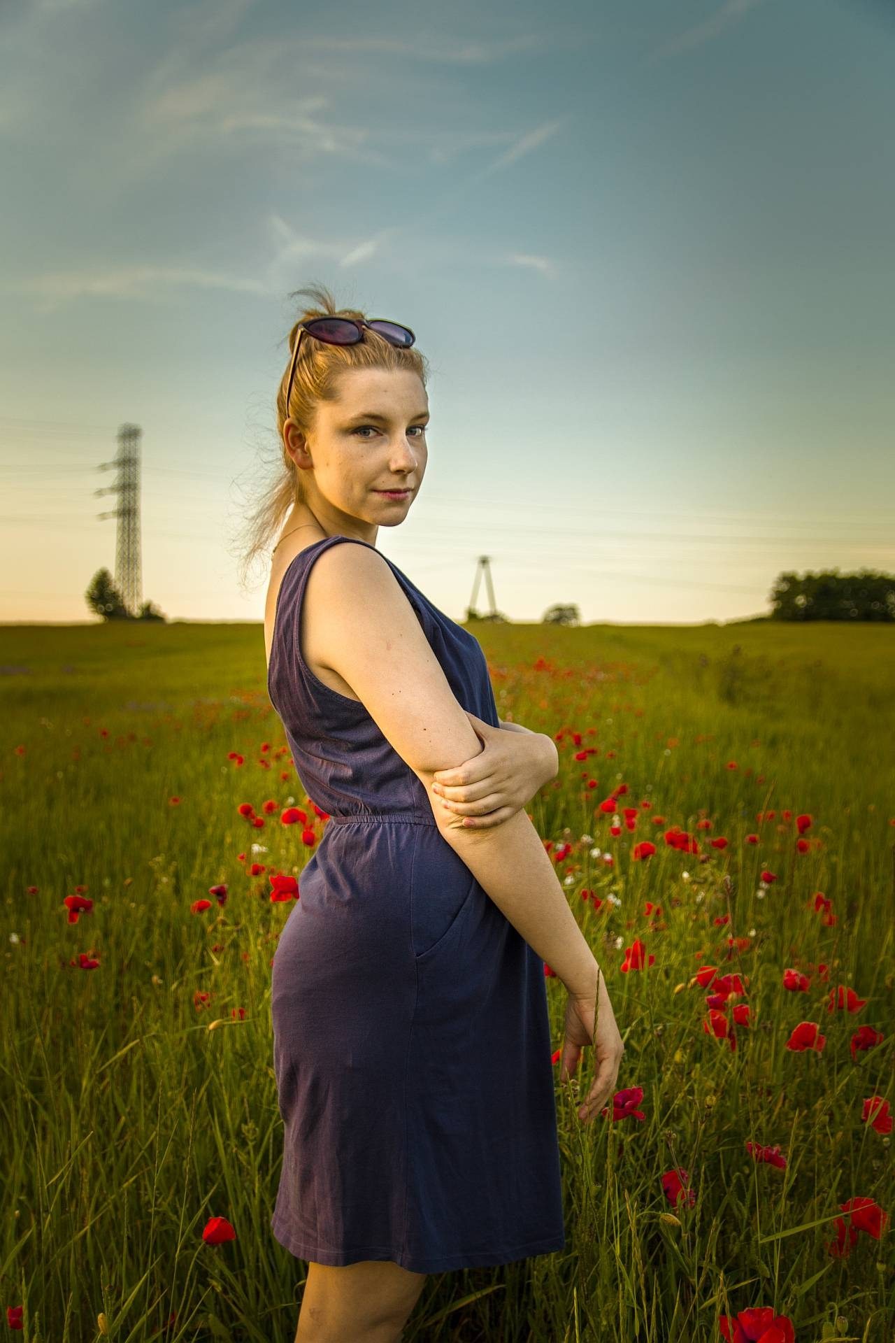 Kobieta w granatowej sukience pozuje na łące z makami, w tle słupy wysokiego napięcia i zachodzące słońce.
