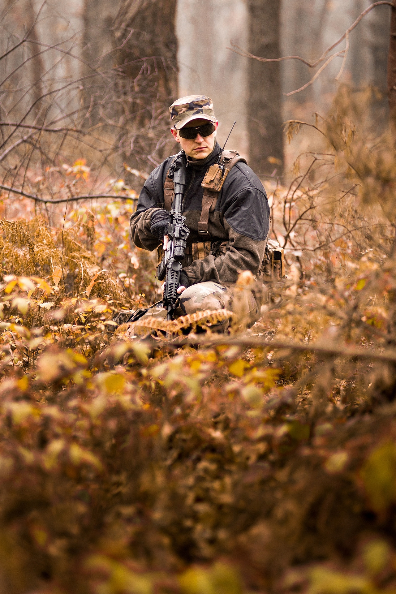 Mężczyzna w kamuflażu klęczy w lesie z karabinem, ubrany w czarną kurtkę i okulary przeciwsłoneczne, wyposażony w radio i taktyczny pas.