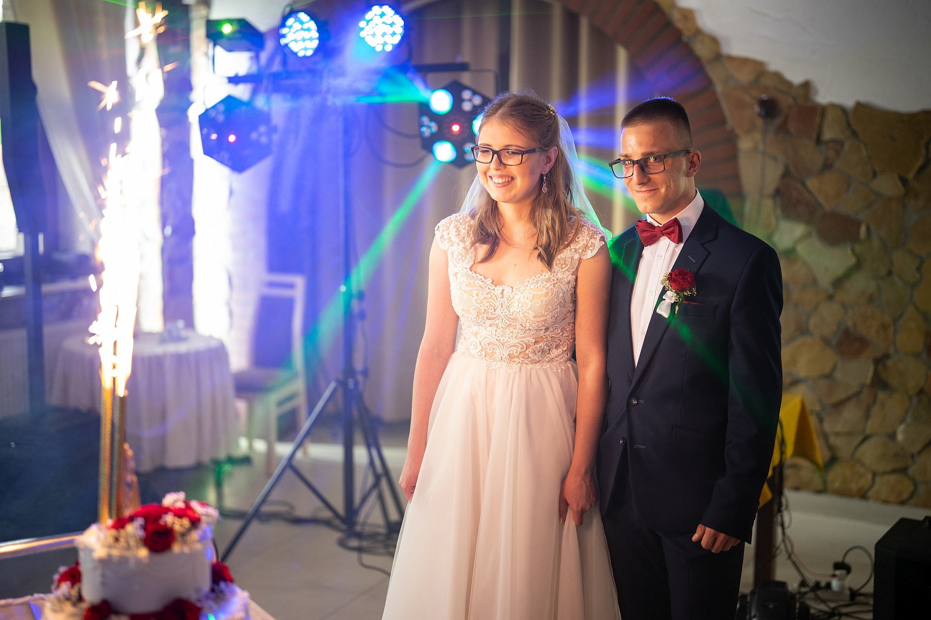 Uśmiechnięta para młoda w okularach stoi obok tortu weselnego z fontannami iskier, oświetlona kolorowymi światłami na tle kamiennej ściany.