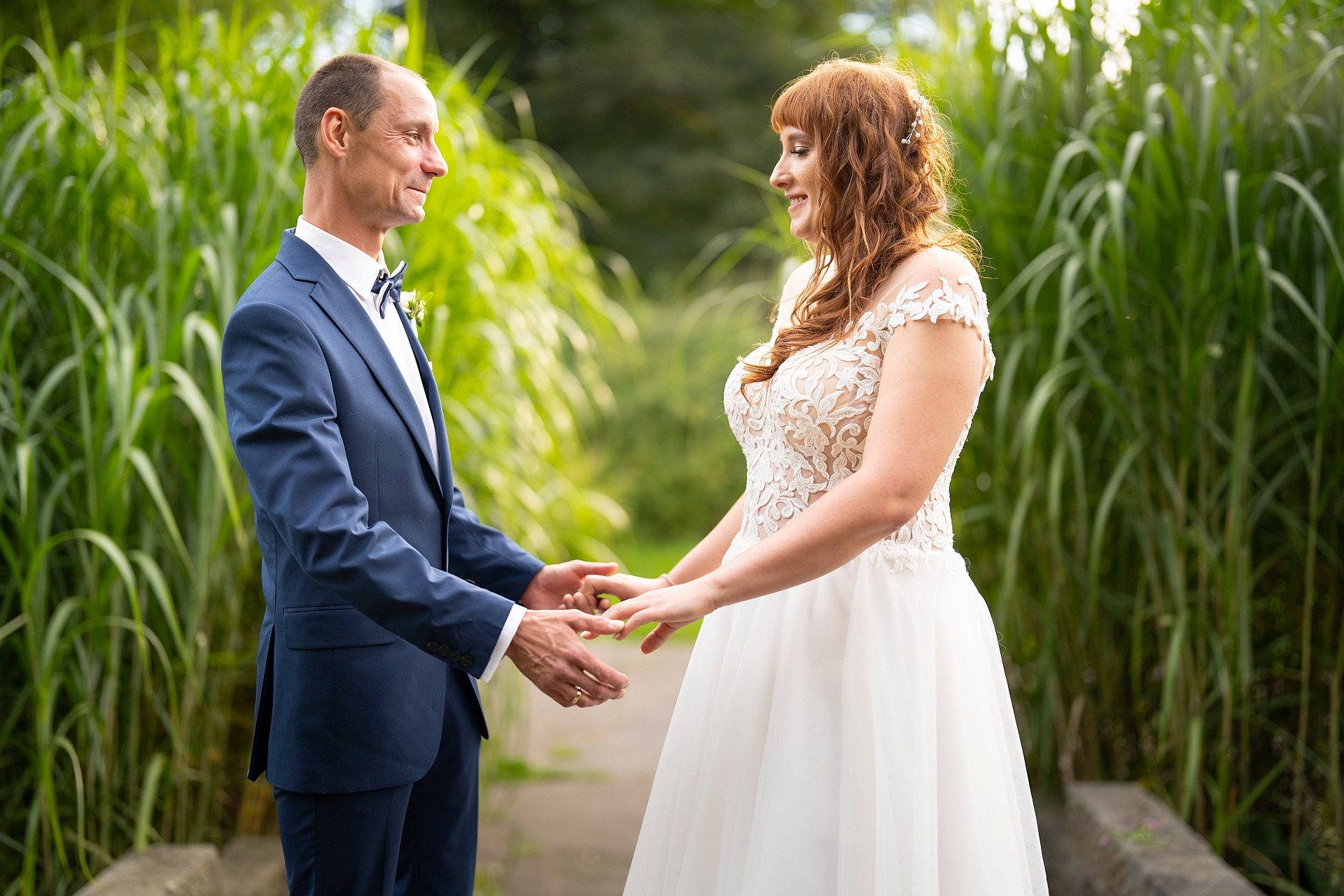 Uśmiechnięta para młoda, ona w koronkowej sukni z odkrytymi ramionami, on w granatowym garniturze z muszką, trzymają się za ręce na tle wysokiej zieleni.