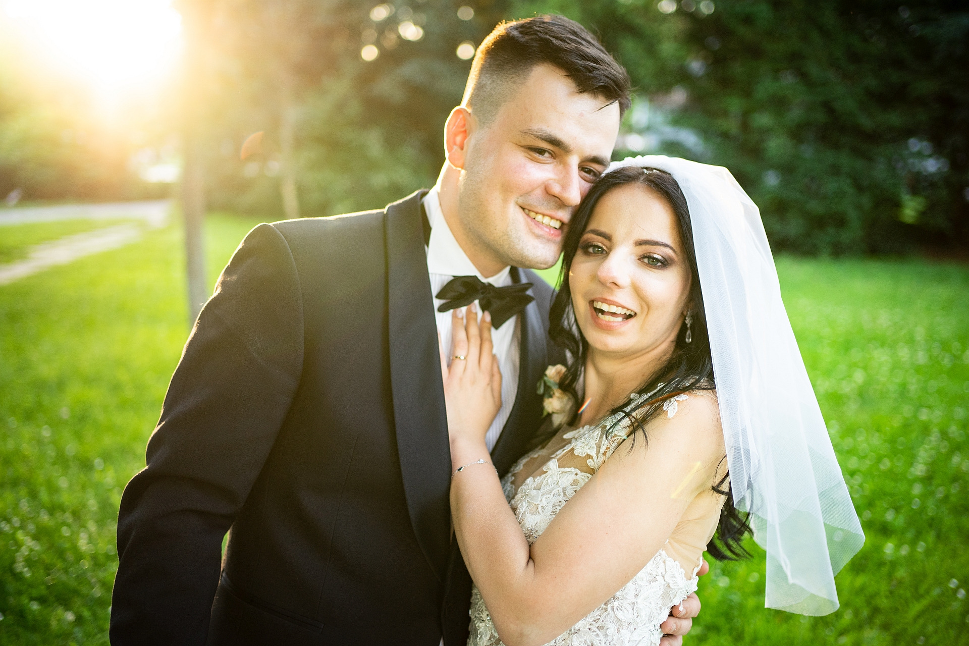 Uśmiechnięta para młoda w Chorzowie, on w czarnym smokingu z muchą, ona w białej sukni ślubnej z welonem, pozują na tle zieleni w słoneczny dzień.
