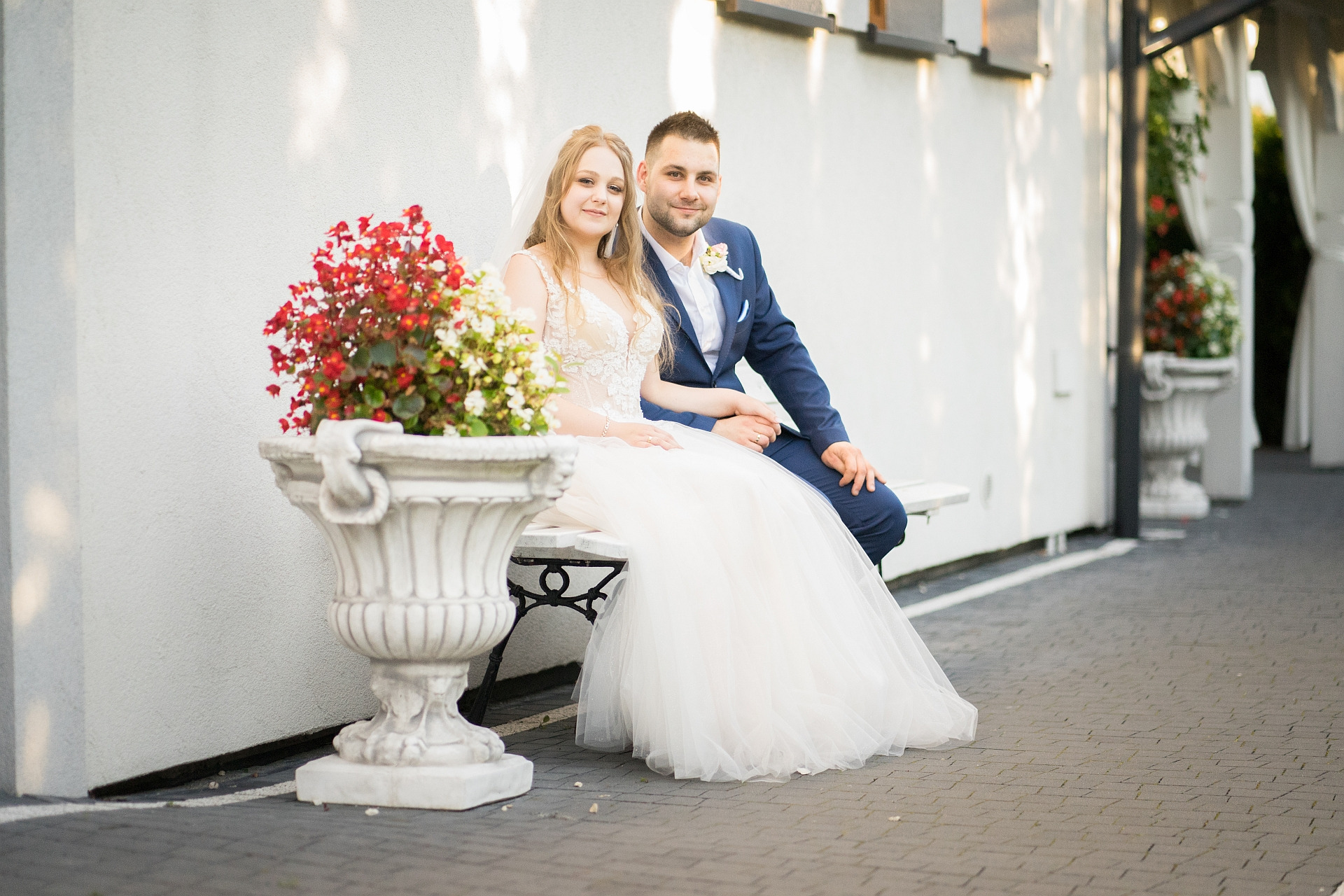 Uśmiechnięta para młoda siedzi na białej ławce obok ozdobnej donicy z czerwono-białymi kwiatami, panna młoda w koronkowej sukni i welonie, pan młody w granatowym garniturze z butonierką.