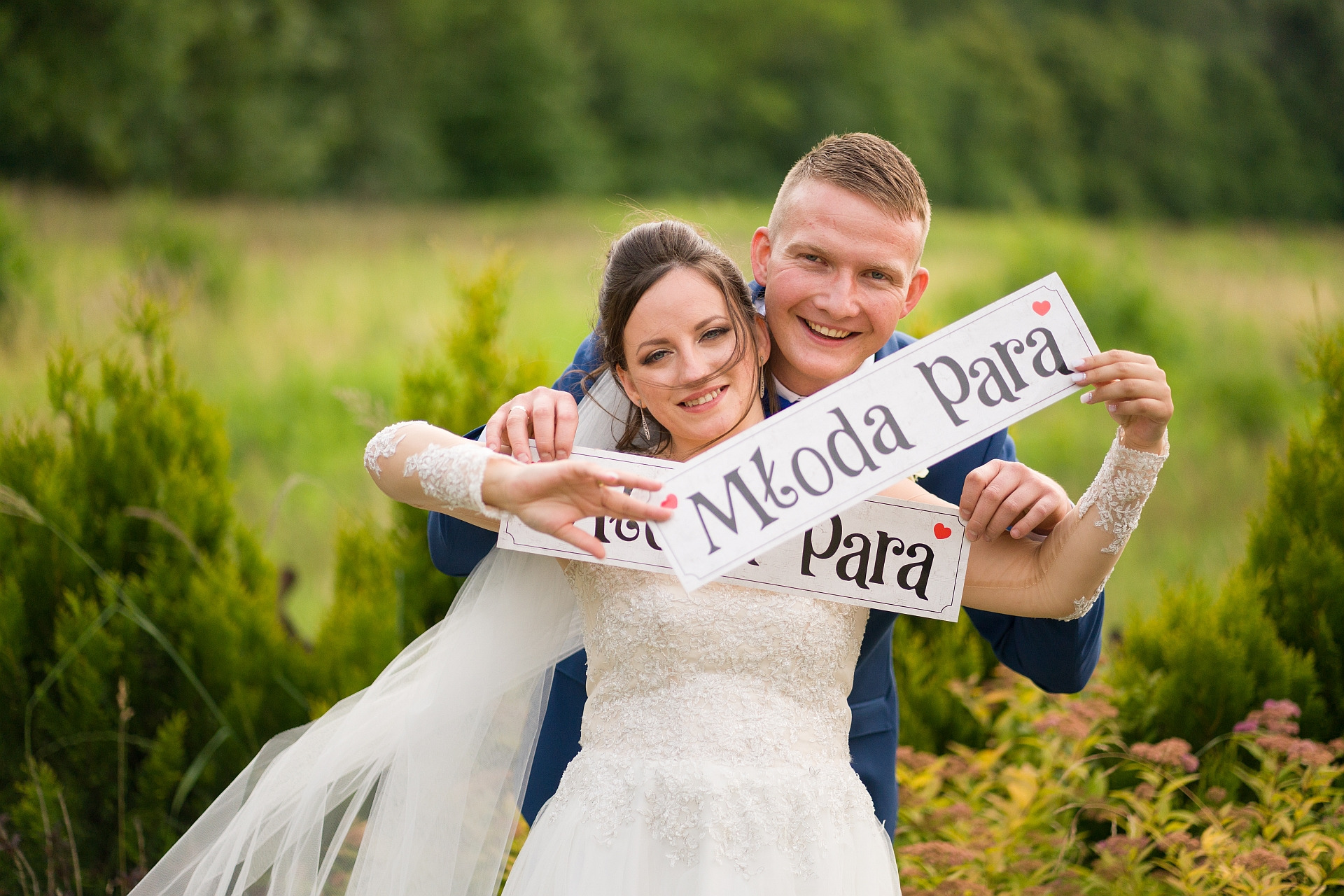 Uśmiechnięta para młoda obejmuje się, trzymając tabliczki z napisem 'Młoda Para' na tle zieleni.