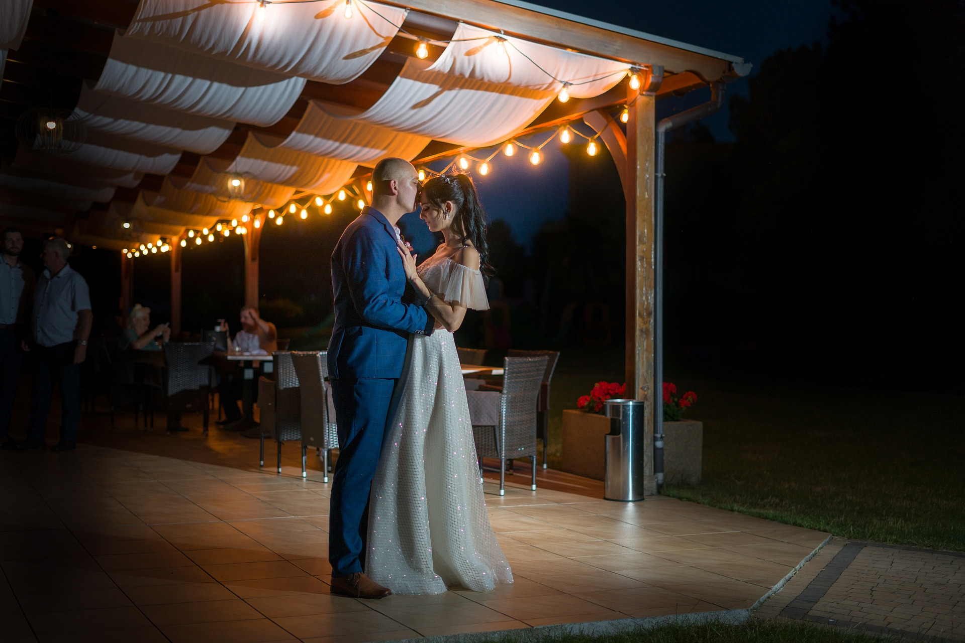 Para młoda tańczy pod drewnianą pergolą oświetloną girlandą żarówek, w tle widoczni goście weselni przy stolikach.