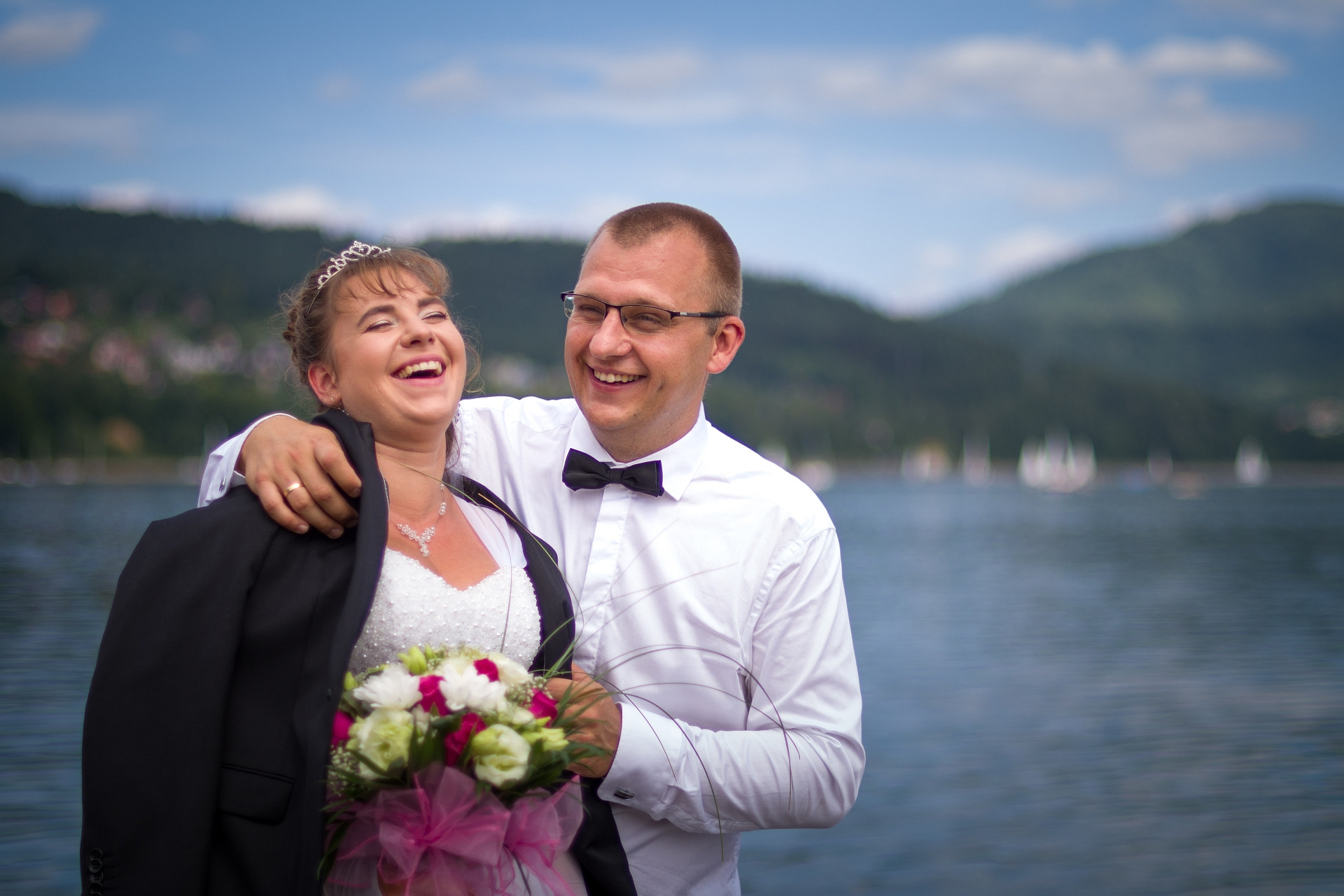 Śmiejąca się para młoda: panna młoda w tiarze i sukni ślubnej z bukietem, pan młody w muszce i koszuli, w tle jezioro i góry.