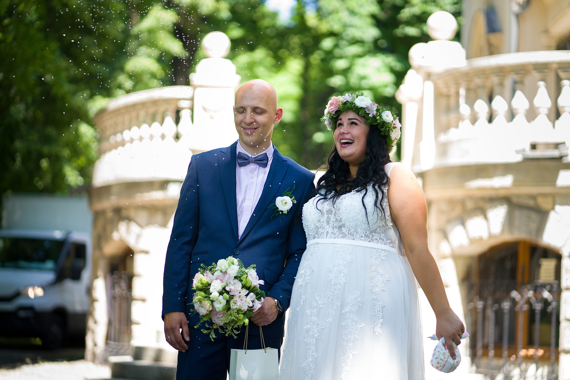 Uśmiechnięta para młoda w plenerze, panna młoda w wianku, pan młody w granatowym garniturze z bukietem, w tle zabytkowa architektura i padający konfetti.