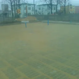 Przygotowany teren pod układanie kostki brukowej, widoczne niebieskie rury wystające z ziemi, w tle budynki przemysłowe i sterta piachu.