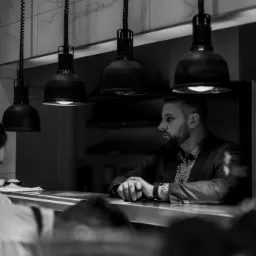 Czarno-białe zdjęcie mężczyzny w garniturze siedzącego za barem w restauracji, z charakterystycznymi lampami wiszącymi nad blatem i marmurowymi płytkami na ścianie w tle.