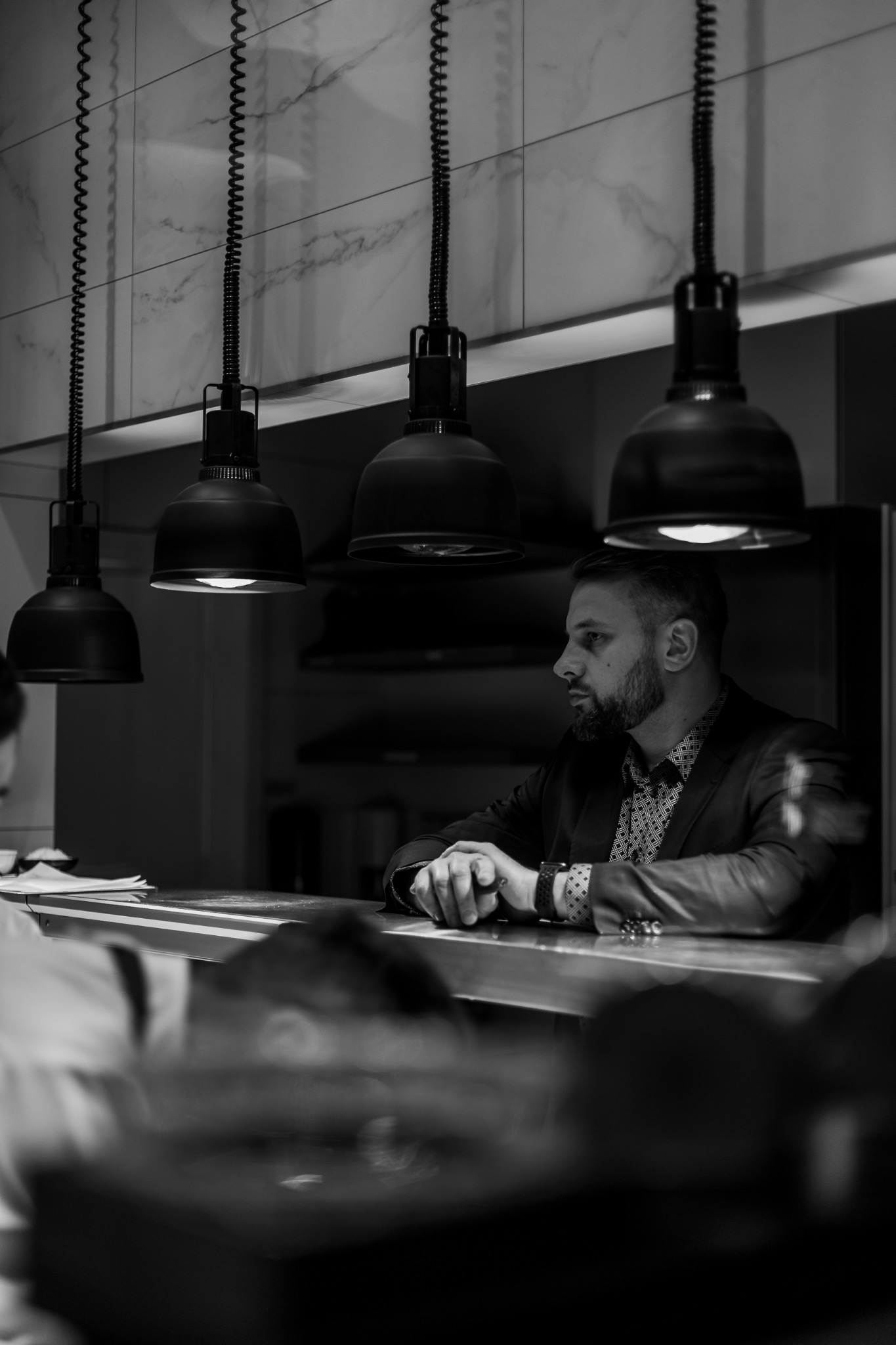 Czarno-białe zdjęcie mężczyzny w garniturze siedzącego za barem w restauracji, z charakterystycznymi lampami wiszącymi nad blatem i marmurowymi płytkami na ścianie w tle.