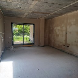 Mateusz p - Surowe wnętrze w trakcie prac wykończeniowych: ściany po tynkowaniu, betonowa posadzka, widoczne przewody elektryczne, okno z widokiem na zieleń.