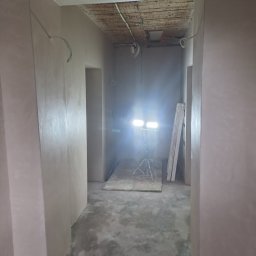 Mateusz p - Wnętrze w trakcie prac wykończeniowych: świeżo otynkowane ściany, widoczne przewody elektryczne, lampa budowlana na statywie, surowy beton na podłodze.