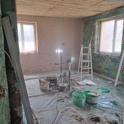 Mateusz p - Pokój w trakcie tynkowania: ściany pokryte świeżym tynkiem, widoczne narzędzia, drabina i lampy oświetleniowe. Podłoga zabezpieczona folią malarską.