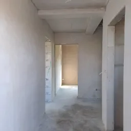 Wnętrze w stanie deweloperskim po tynkowaniu maszynowym, widoczne gładkie ściany, surowy betonowy strop z wystającymi kablami i otwory drzwiowe.