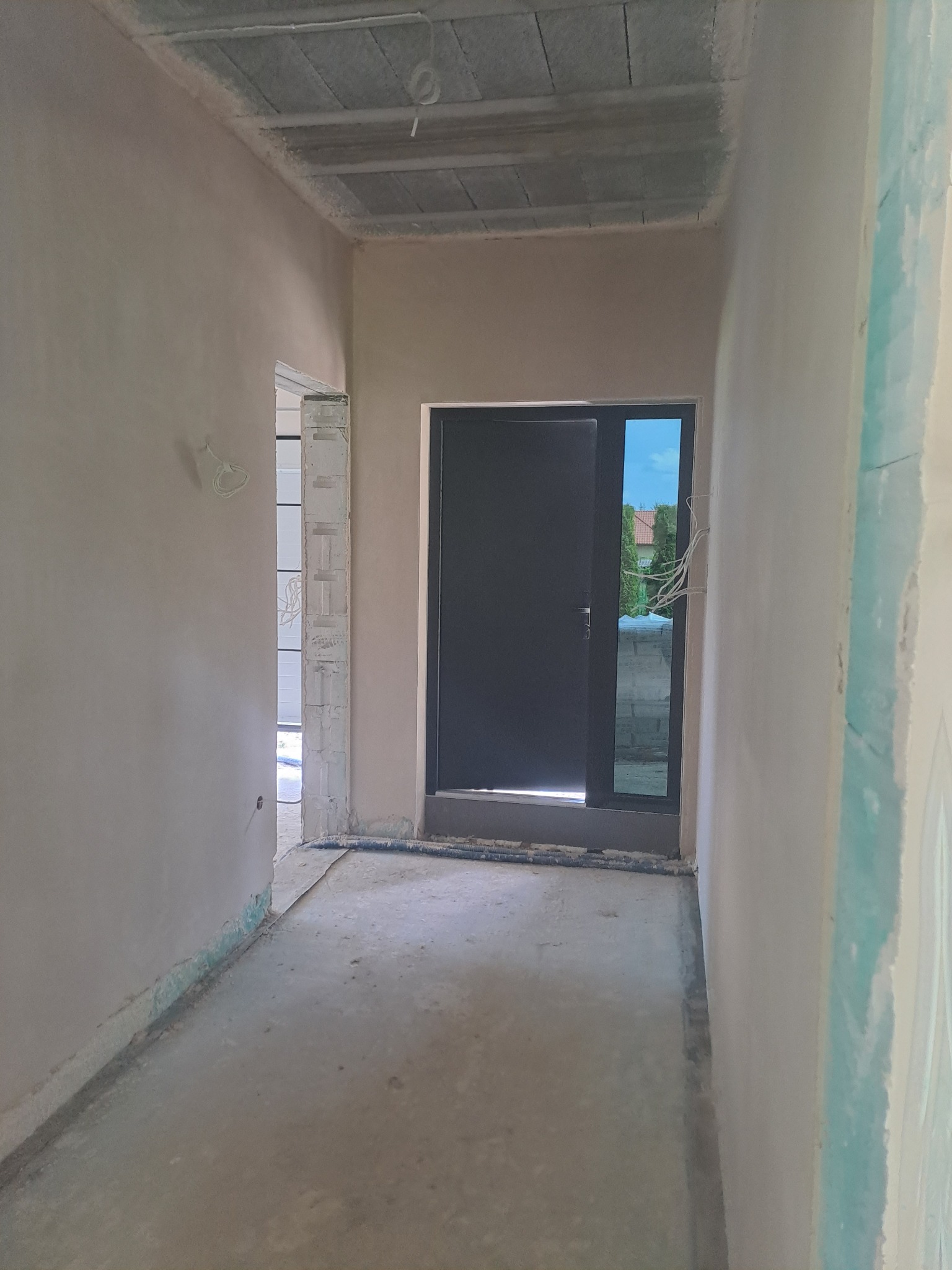 Wnętrze w trakcie prac wykończeniowych: ściany pokryte tynkiem, widoczne przewody elektryczne przy ościeżnicy, surowy strop i podłoga. W tle widok na zieleń przez okno.