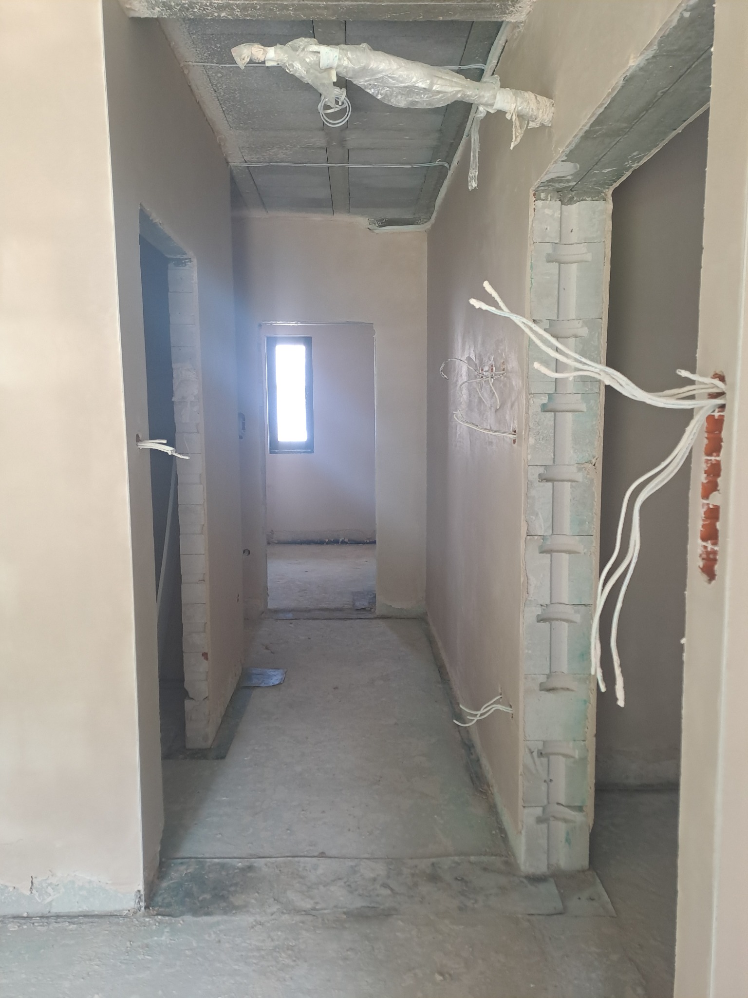 Wnętrze w trakcie prac wykończeniowych: świeżo otynkowane ściany, widoczne przewody elektryczne, surowy betonowy sufit z instalacją zabezpieczoną folią. W tle okno.