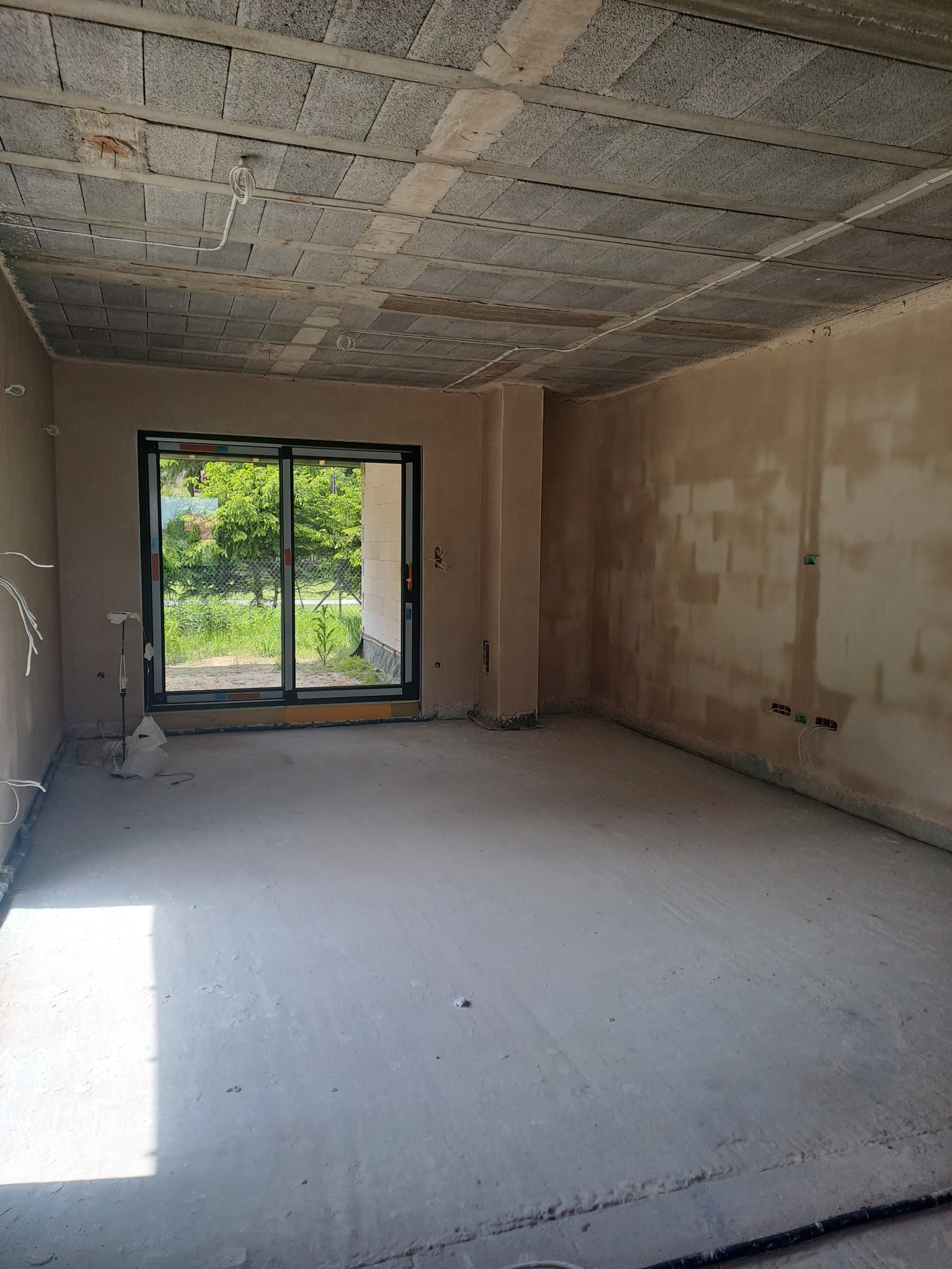 Surowe wnętrze w trakcie prac wykończeniowych: ściany po tynkowaniu, betonowa posadzka, widoczne przewody elektryczne, okno z widokiem na zieleń.