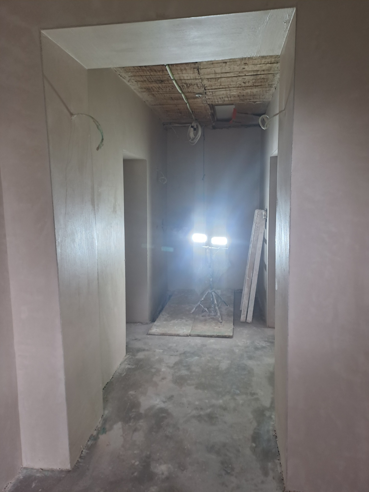 Wnętrze w trakcie prac wykończeniowych: świeżo otynkowane ściany, widoczne przewody elektryczne, lampa budowlana na statywie, surowy beton na podłodze.