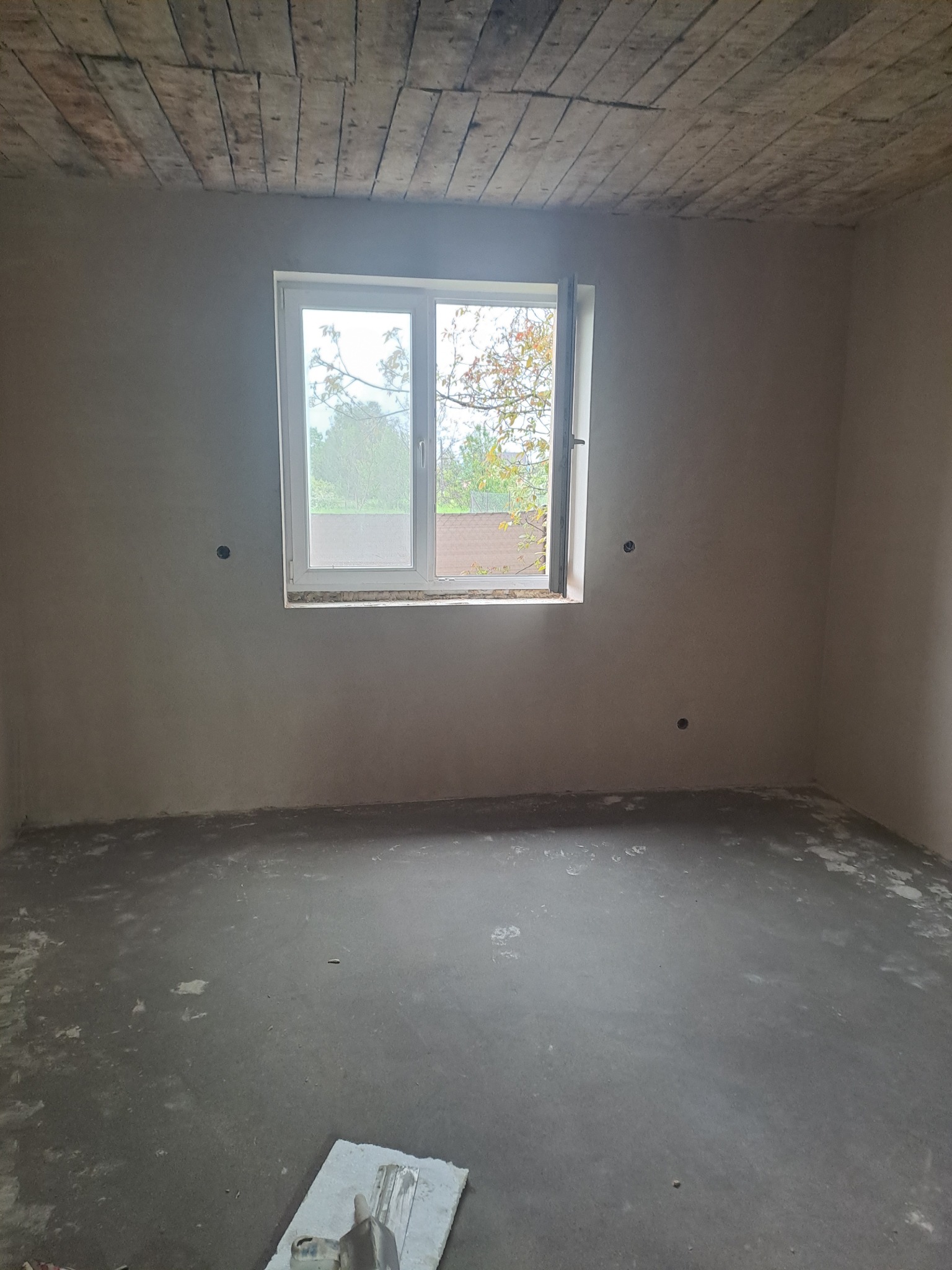 Surowe wnętrze pokoju po tynkowaniu ścian, z widocznym oknem i narzędziami na podłodze. Sufit z drewnianych desek dodaje rustykalnego charakteru.