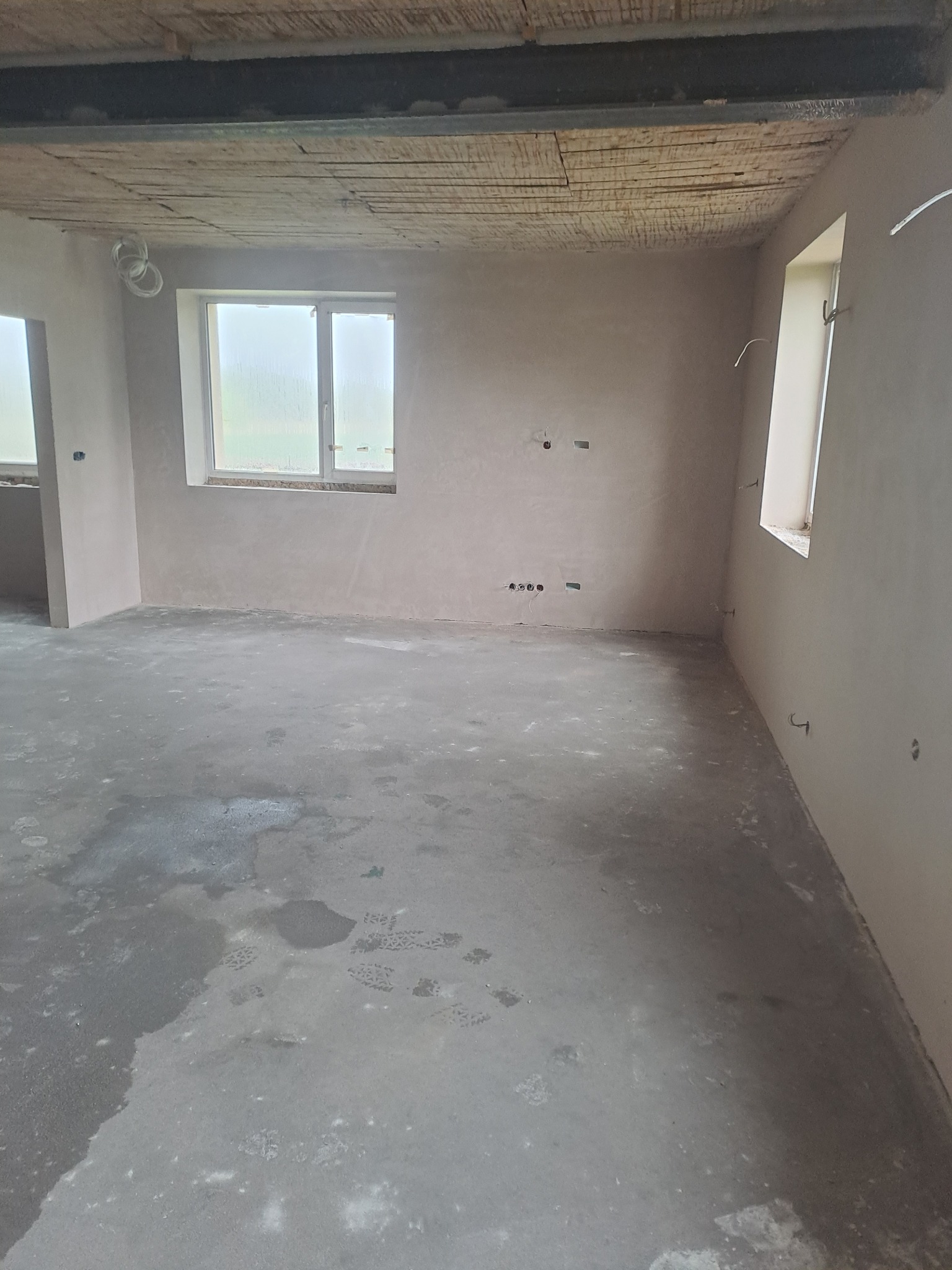 Wnętrze po tynkowaniu maszynowym: ściany pokryte gładzią, widoczne okna, surowy betonowy sufit i mokra posadzka z odciskami butów.