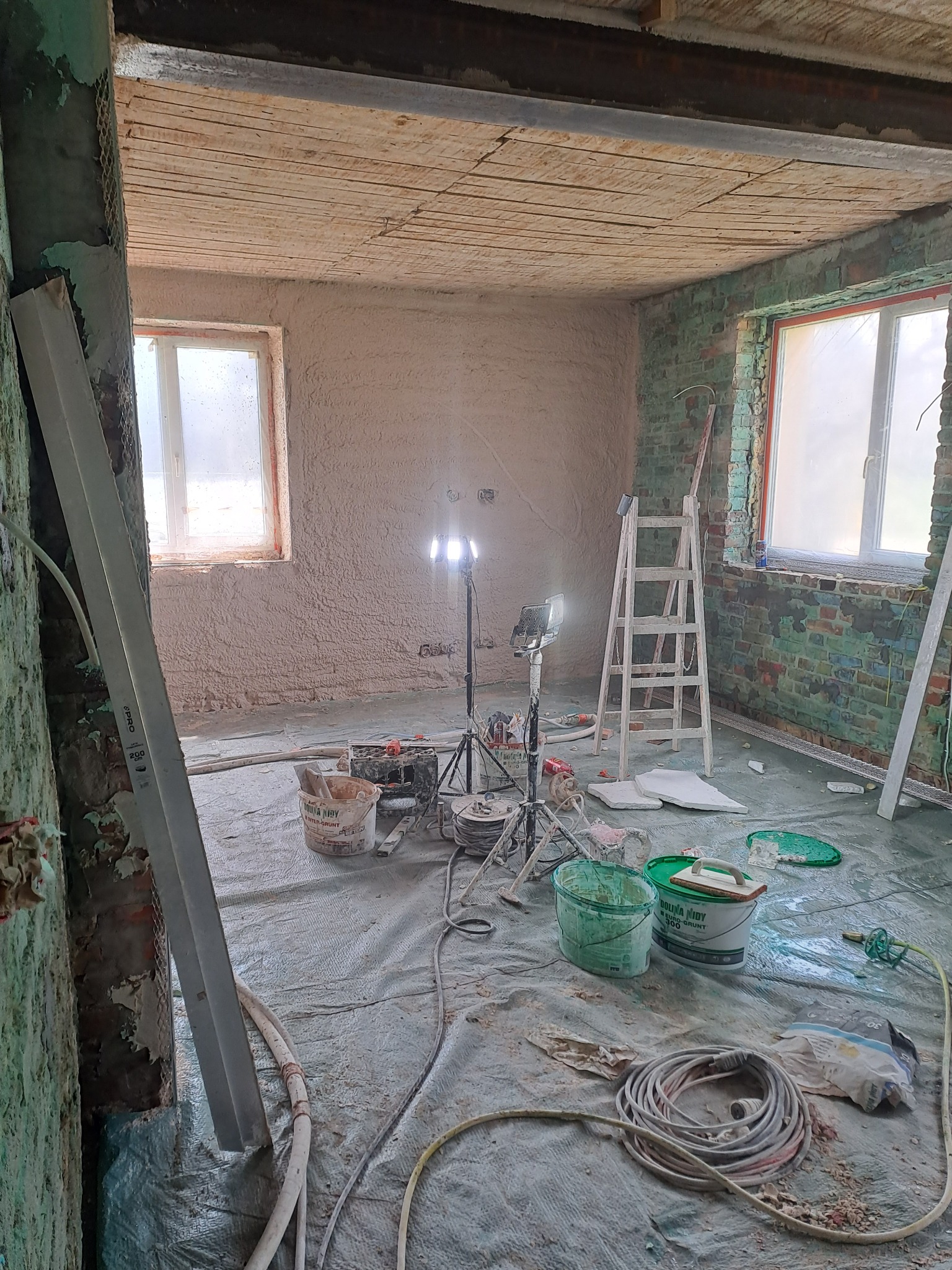 Pokój w trakcie tynkowania: ściany pokryte świeżym tynkiem, widoczne narzędzia, drabina i lampy oświetleniowe. Podłoga zabezpieczona folią malarską.