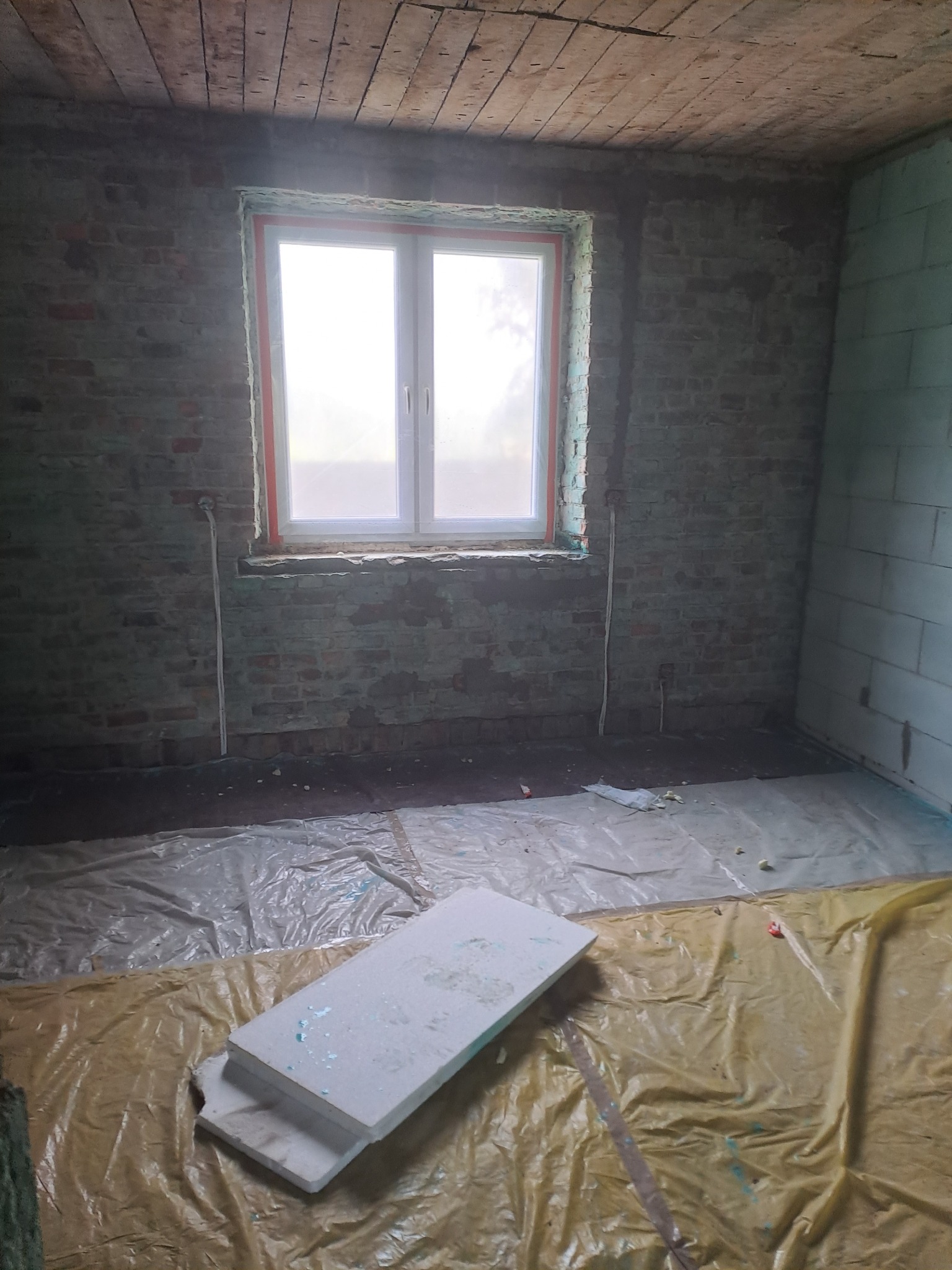 Wnętrze pokoju w trakcie remontu: surowe cegły, nowe okno z pomarańczowym uszczelnieniem, izolacja podłogi folią i styropianem. Widoczne ślady wcześniejszych prac.