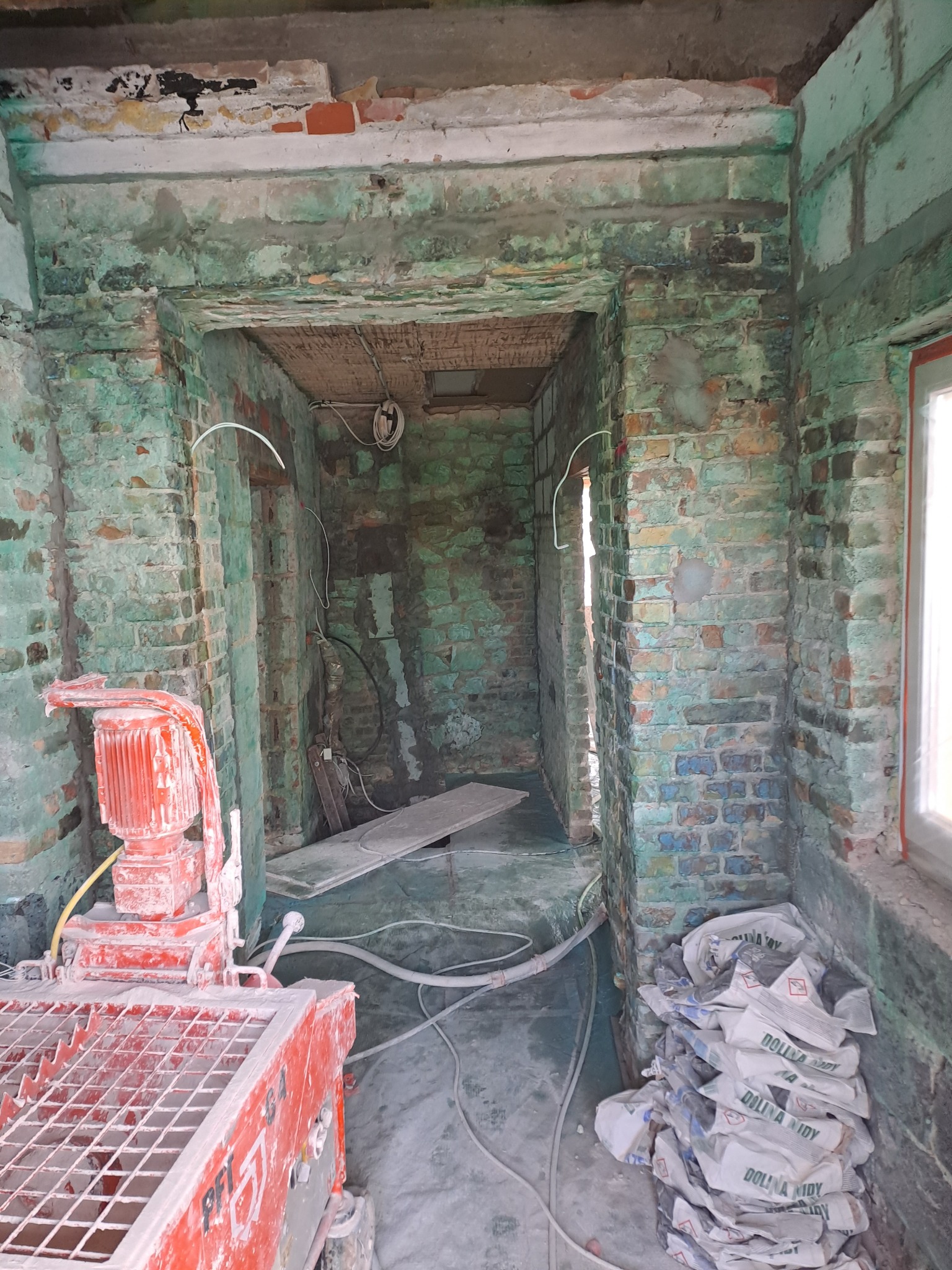 Wnętrze budynku w trakcie remontu. Widoczna maszyna do tynkowania, worki z zaprawą i fragmenty odsłoniętej cegły. Ściany z resztkami farby w odcieniach zieleni i błękitu.