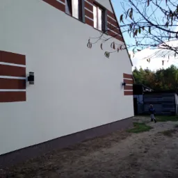 Ściana domu z ociepleniem i dekoracyjnymi brązowymi pasami, oświetlenie zewnętrzne, fragment okna, w tle budynki gospodarcze i dziecko na podwórku.