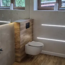 Nowoczesna łazienka po remoncie: podwieszana toaleta z drewnianą obudową, drewnopodobna podłoga, ściana z dekoracyjnym tynkiem i oświetleniem LED, okno z widokiem na zieleń.