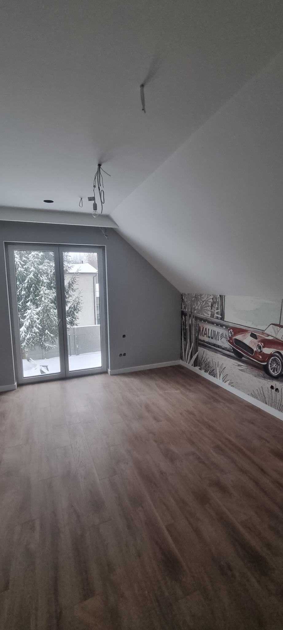 Poddasze po remoncie z drewnianą podłogą, szarą ścianą i tapetą z motywem czerwonego samochodu, widok na zaśnieżone drzewa przez okno balkonowe, widoczne niedokończone instalacje elektryczne.