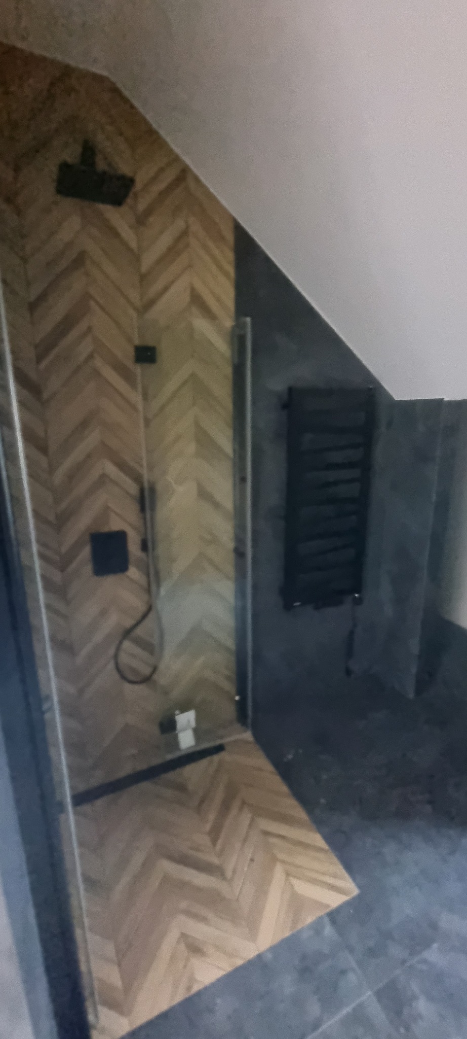 Ukończona kabina prysznicowa w łazience na poddaszu, z wykończeniem ścian w jodełkę z jasnego drewna i ciemnymi płytkami na podłodze oraz grzejnikiem drabinkowym.