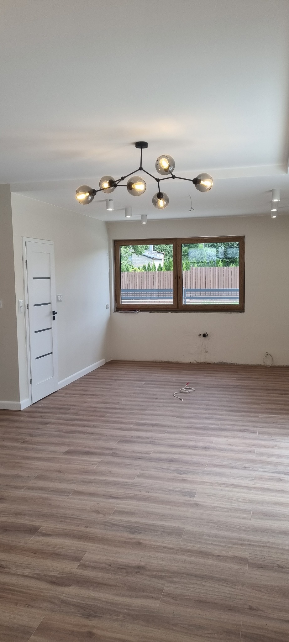 Wnętrze pokoju po pracach wykończeniowych: ułożona podłoga z paneli imitujących drewno, pomalowane ściany na biało, zamontowane drzwi i okno z drewnianą ramą, nowoczesna lampa sufitowa.