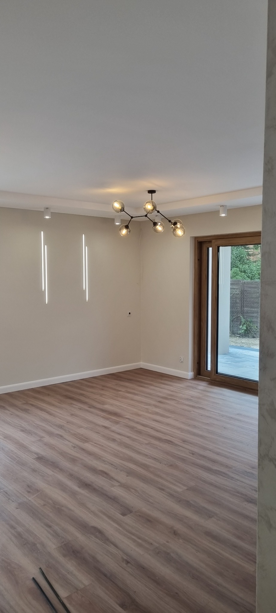 Nowoczesne wnętrze po remoncie z drewnopodobną podłogą, designerską lampą sufitową i pionowymi lampami ściennymi, widoczne listwy przypodłogowe i drzwi przesuwne.