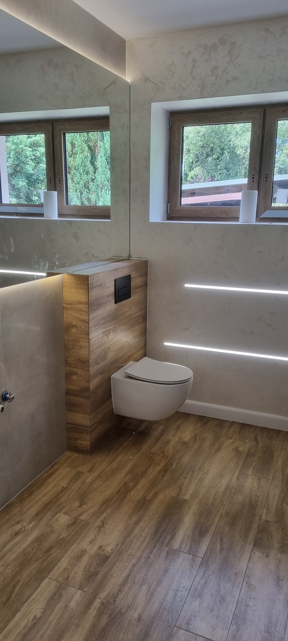 Nowoczesna łazienka po remoncie: podwieszana toaleta z drewnianą obudową, drewnopodobna podłoga, ściana z dekoracyjnym tynkiem i oświetleniem LED, okno z widokiem na zieleń.