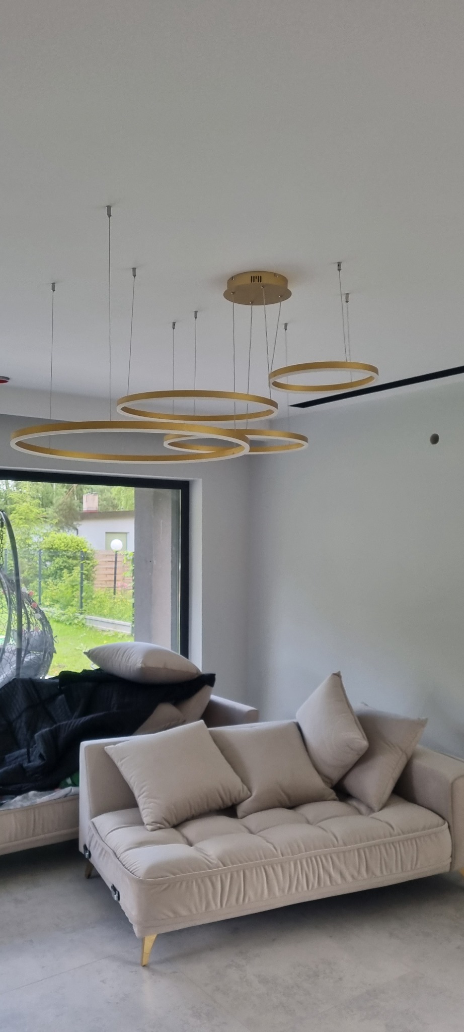 Nowoczesny salon z beżową sofą i designerskim oświetleniem w postaci złotych okręgów, widok na ogród przez duże okno.